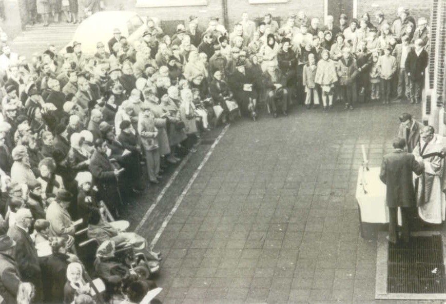 Een Heilige mis op het kerkplein. Een kerkdienst buiten voor St.Hippolytuskerk. Wim staat in de menigte tegen de muur rechts. Zijn zoon Wim is misdienaar rechts van de priester